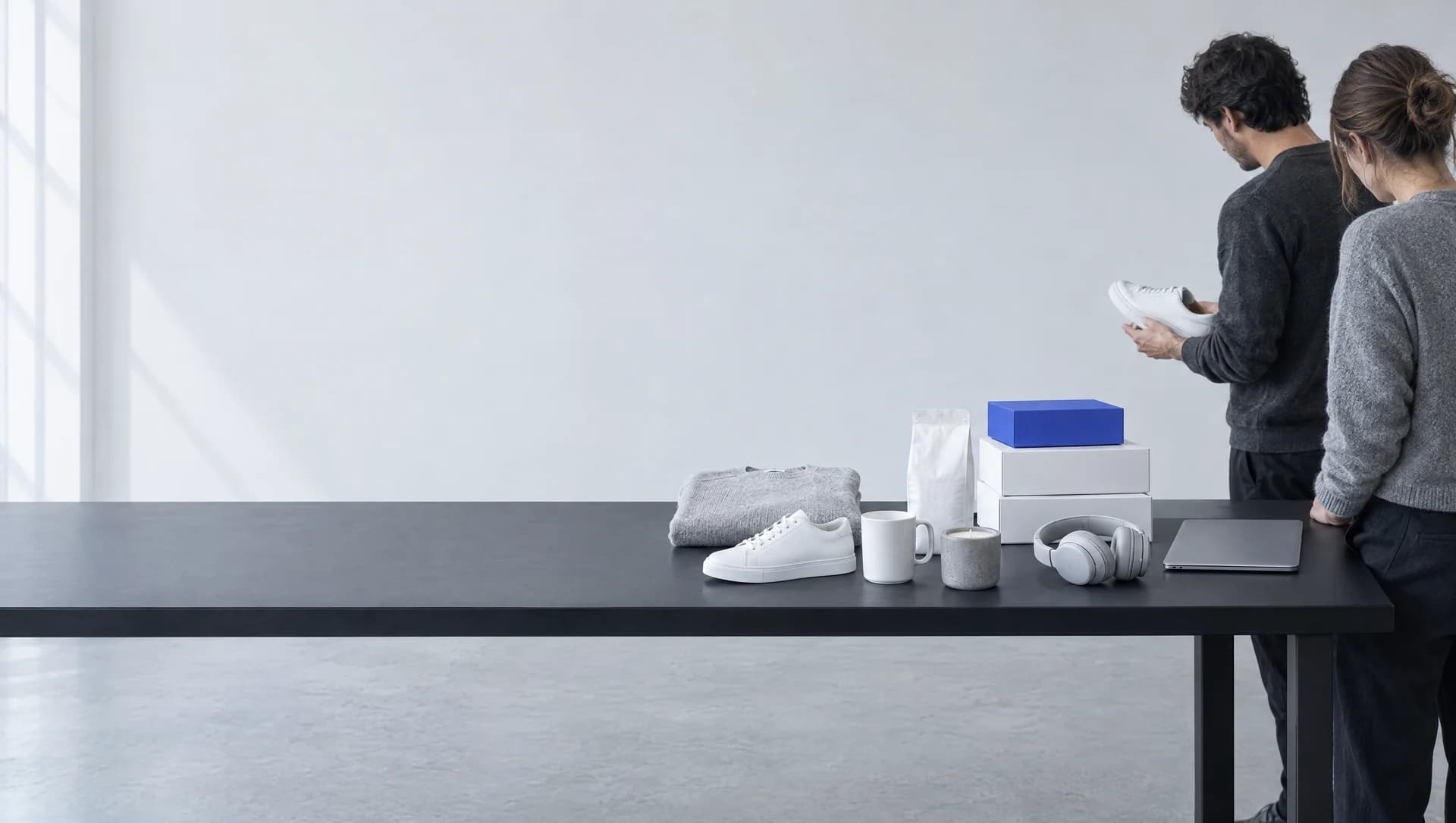 A long black table in a pale, empty room. Unbranded samples line its right half: a folded gray knit, a white sneaker, a mug, a candle, a coffee bag, headphones, a closed laptop and two white boxes topped by a blue one. At the right end, two people stand looking at a white sneaker one of them holds.