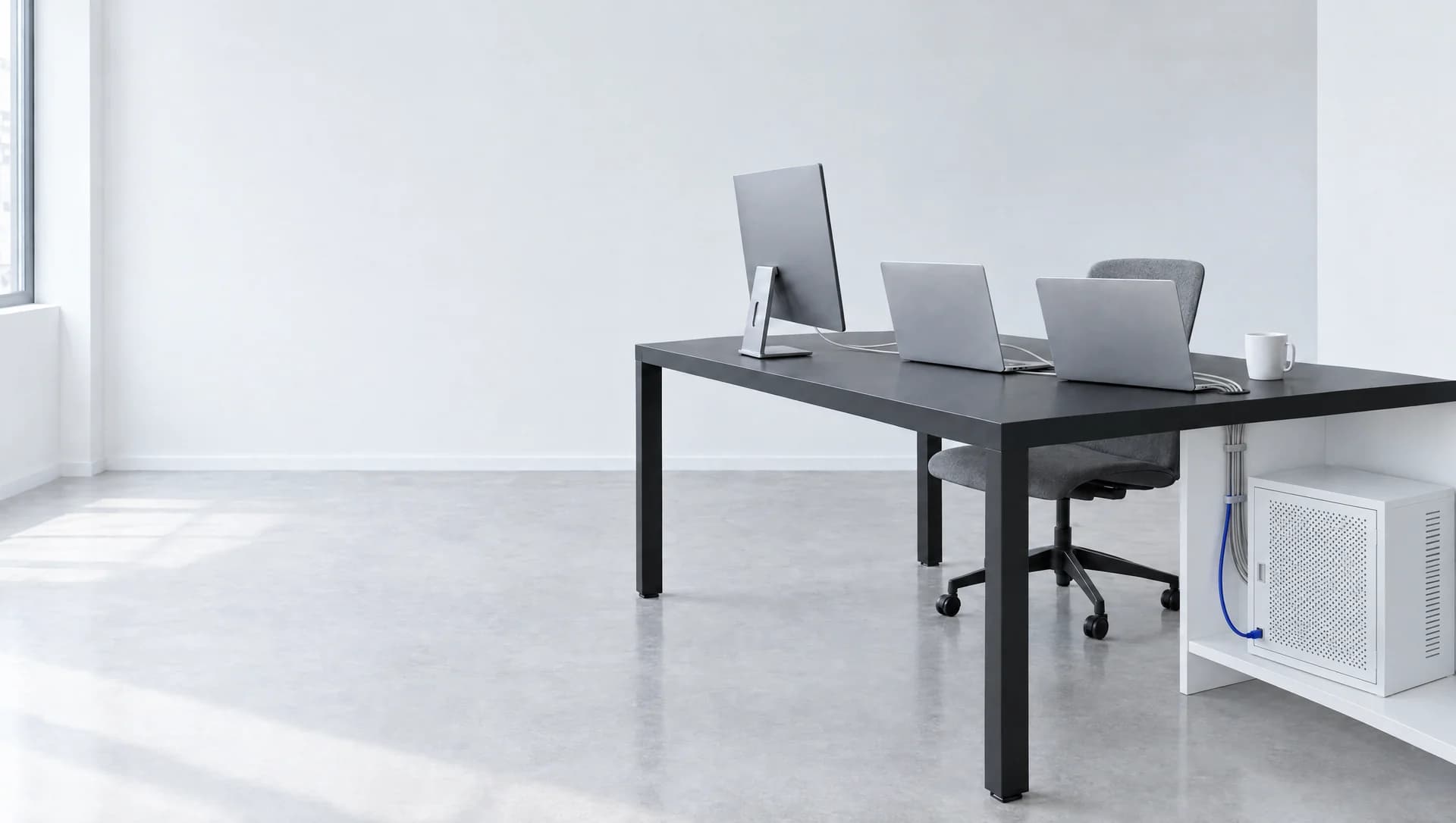 A bright white room in daylight. A black desk on the right holds a monitor and two open laptops, all turned away from the camera, and a white mug, with a gray chair tucked in behind. Under the desk, a white vented network cabinet sits on a low shelf, fed by a bundle of gray cables and one blue cable. The left side is open wall and concrete floor.