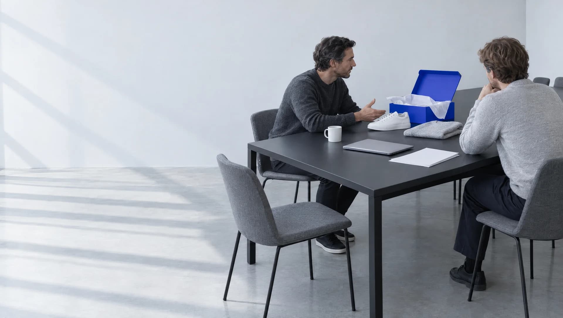 Two men in gray sweaters sit across the corner of a long black table in a bright room, mid-conversation. One gestures toward a white sneaker next to an open blue box lined with white tissue. A closed laptop, a folded knit, a mug and blank paper lie on the table, and an empty chair is pulled out in the foreground.
